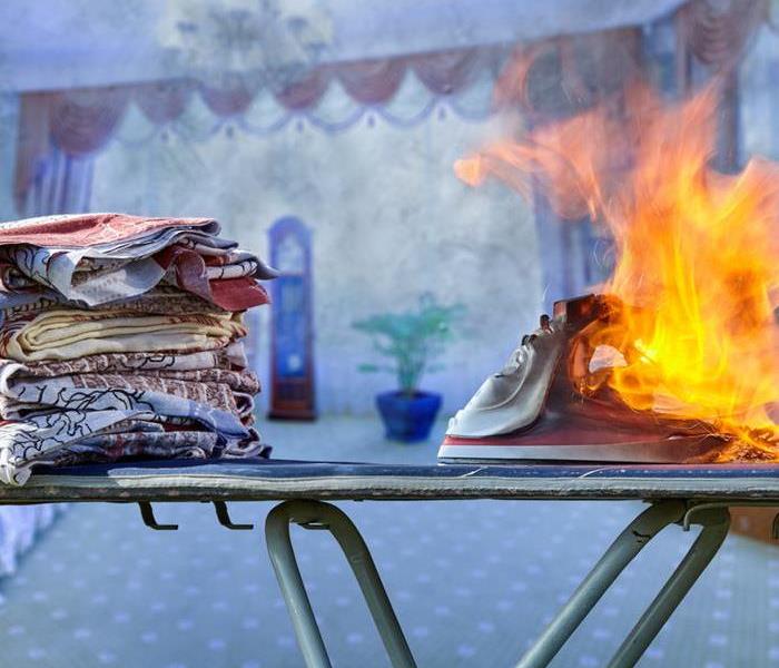 An iron has melted and is covered in flames as it rests on an ironing board in a high-end looking bedroom