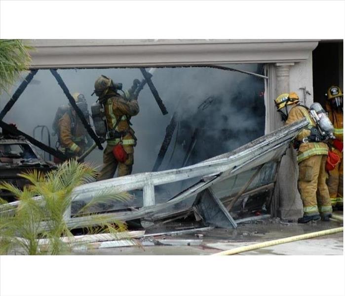 Firemen use hoses to extinguish a fire in a house garage. A melted garage door and burnt car shows high heat and damages