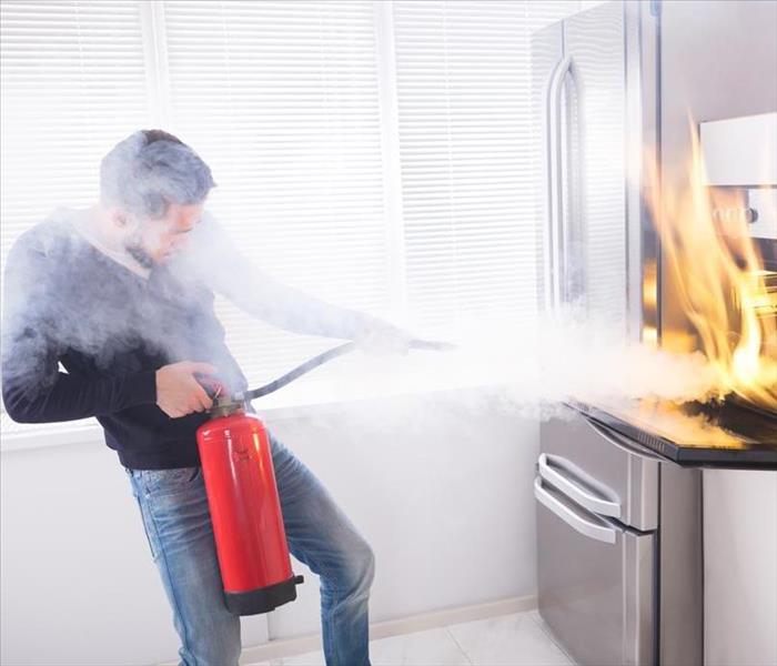 A man uses all his might to properly use a fire extinguisher by aiming at the base of the flames coming from the oven