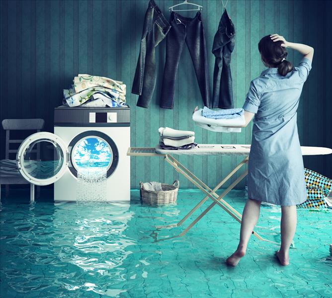 A laundry room shows room flooding out from a running clothes washing machine as a barefoot lady stands there was ironing but