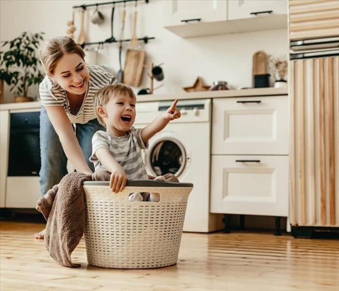 A toddler smiles big as he sits inside a laundry basket being pushed by his mom in a fresh clean looking kitchen