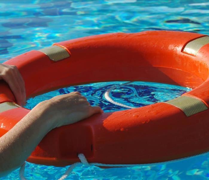 A turquoise blue pool of water has a bright orange safety floatation device with a rope. Young hands are holding on 