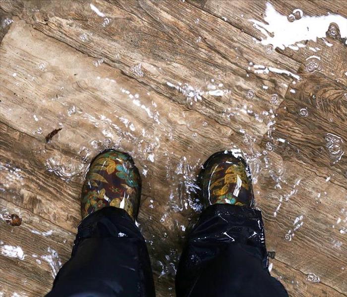 Two feet wearing water proof galoshes, stepping on a wood floor that has been flooded and covered in water 