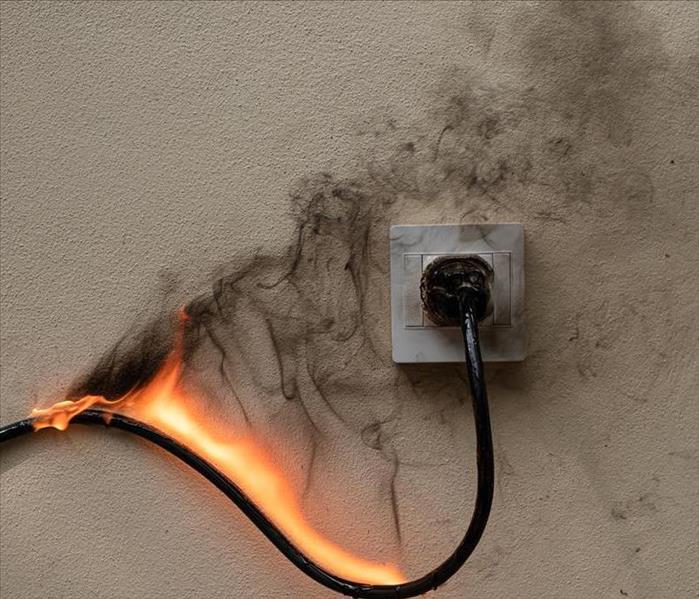 A wall outlet has a full sized plug. The chord of it is on fire and black smoke is beginning to cover the wall