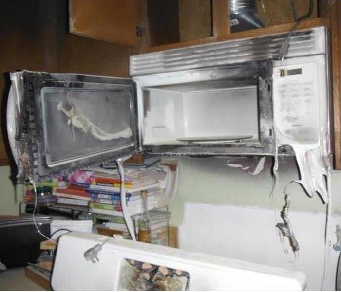 A microwave above a stove has been affected by heat and smoke. The door and front panel are melted and dripping